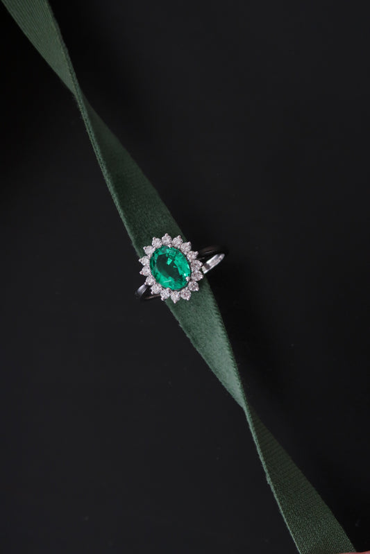 Sunburst Halo Ring in Emerald — vivid green oval emerald centre with round diamond halo, editorial shot on dark background with green velvet ribbon — Stone Symphony