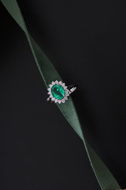 Sunburst Halo Ring in Emerald — vivid green oval emerald centre with round diamond halo, editorial shot on dark background with green velvet ribbon — Stone Symphony