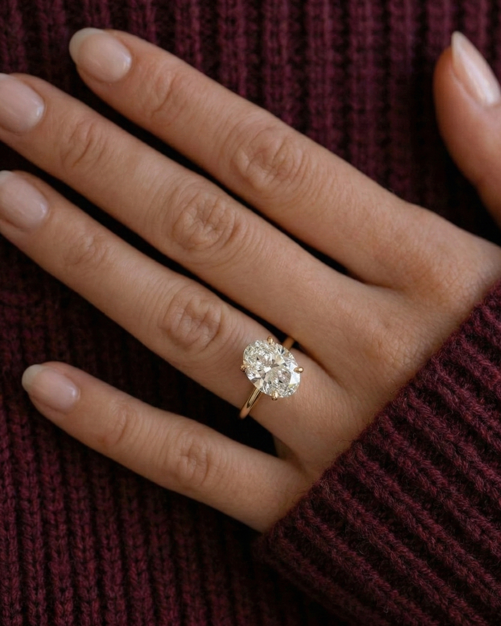 Oval Solitaire Diamond Ring on finger — yellow gold solitaire, maroon textured background — Stone Symphony